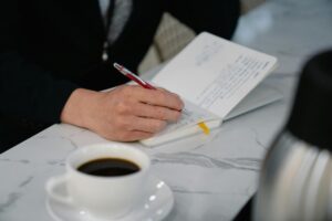 A faculty member sitting at a desk writing, with an open notebook and coffee, planning summer research goals